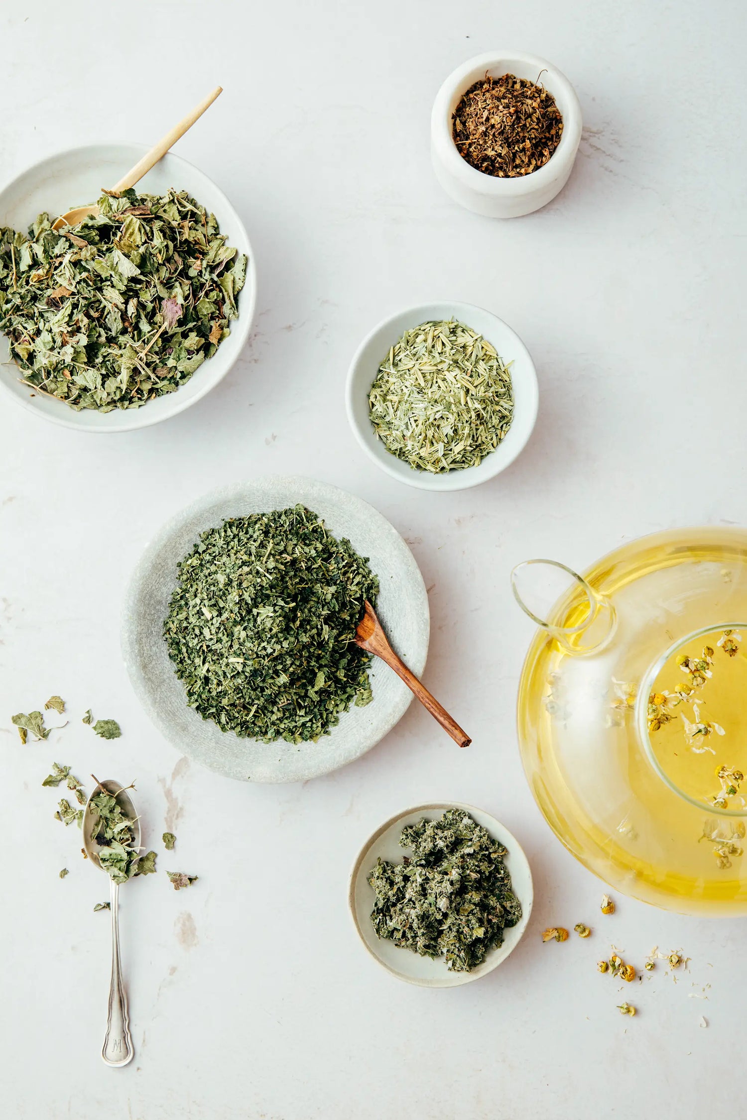 Bowls of various sizes filled with various green and red herbs next to a glass teapot filled with a yellow elixir